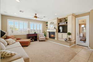 Living area with a glass covered fireplace, a ceiling fan, light tile patterned floors, light carpet, and built in features