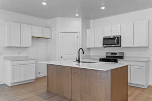 Kitchen featuring an island with sink, stainless steel appliances, white cabinetry, light wood-style flooring, and light stone countertops