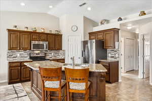 Kitchen featuring stainless steel appliances, a center island with sink, light stone countertops, a kitchen bar, and vaulted ceiling