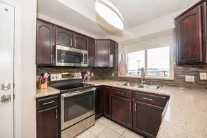 Kitchen featuring stainless steel appliances, dark wood finish cabinets, light tile patterned floors, decorative backsplash, and light stone counters