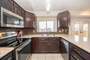 Kitchen featuring stainless steel appliances, dark wood finish cabinets, tasteful backsplash, light tile patterned flooring, and light stone counters