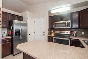 Kitchen with dark wood finish cabinetry, stainless steel appliances, lofted ceiling, light tile patterned floors, and backsplash