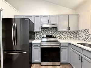 Kitchen with gray cabinetry, freestanding refrigerator, stainless steel electric stove, vaulted ceiling, and light stone counters