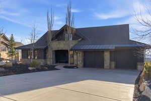 View of front facade with a standing seam roof, stone siding, driveway, a shingled roof, and an attached garage