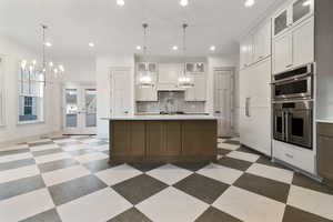 Two tone kitchen with glass fronted cabinets, hanging lights, stainless steel double oven, two tone cabinets, and an island with sink