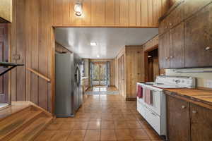 Kitchen with white electric range, stainless steel fridge with ice dispenser, butcher block counters, dark wood finish cabinets, and wooden walls