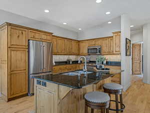 Kitchen featuring dark stone countertops, stainless steel appliances, an island with sink, a kitchen bar, and light wood finished floors