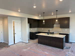 Kitchen featuring dark wood finish cabinetry, a center island with sink, hanging light fixtures, tasteful backsplash, and light stone countertops