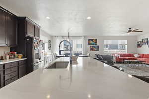Kitchen featuring dark wood finish cabinets, stainless steel refrigerator with ice dispenser, light quartz countertops, open floor plan, and ceiling fan