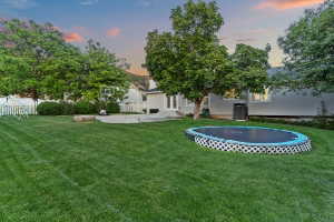 Expansive backyard featuring a large green lawn, mature trees, a concrete patio, and a white picket fence