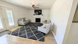Spacious living area featuring light wood-finish flooring, a white mantel fireplace, and a ceiling fan with integrated lighting