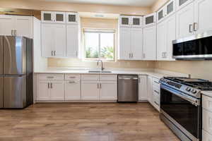 Kitchen with glass insert cabinets, stainless steel appliances, white cabinetry, light wood finished floors, and light stone counters