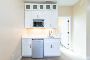 Kitchen featuring white cabinetry, white dishwasher, stainless steel microwave, glass fronted cabinets, and light stone counters