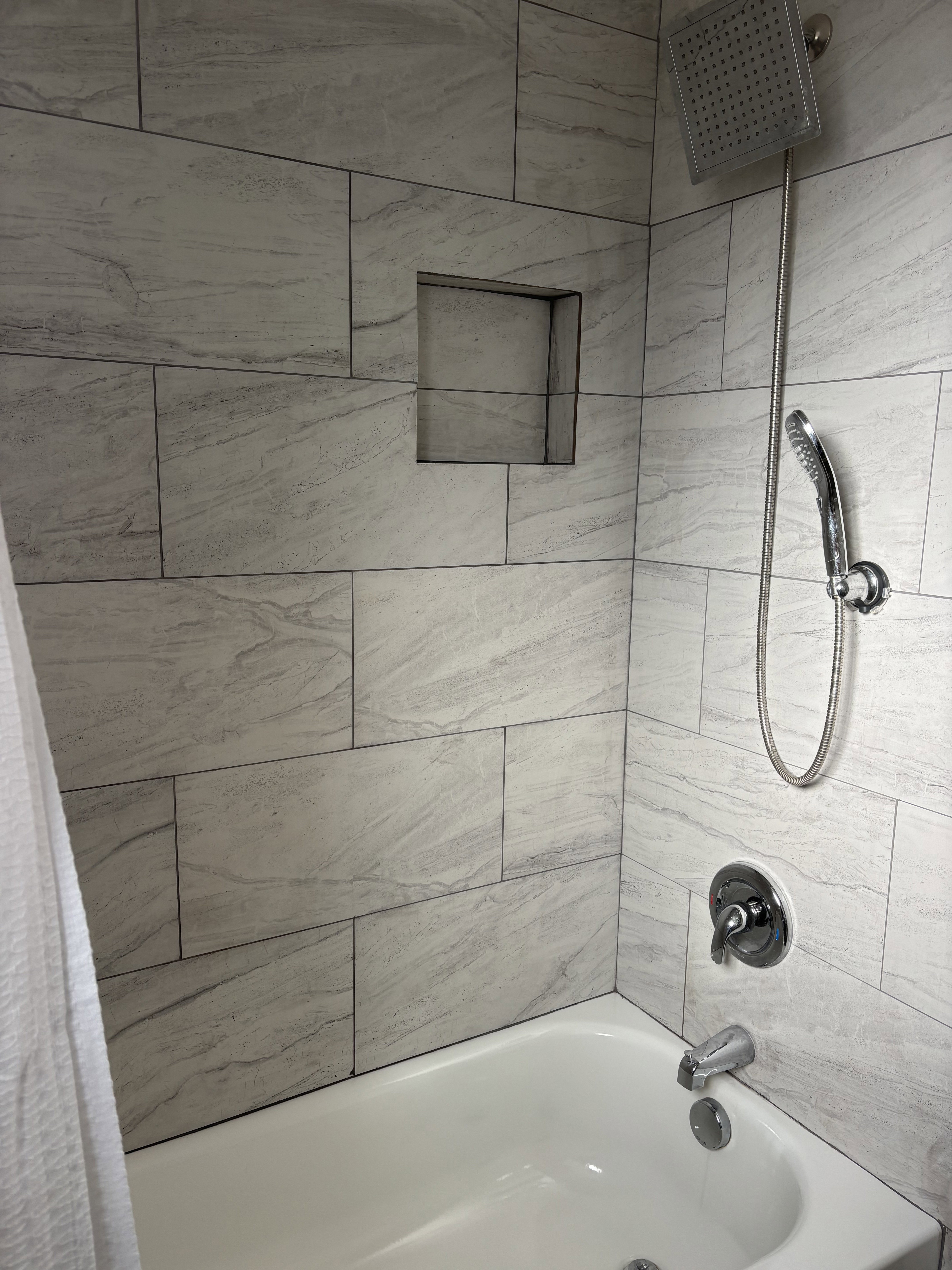 Bathtub and shower combination featuring large format grey-toned tile, a recessed shower niche, a rain showerhead, a handheld shower fixture, and polished chrome hardware