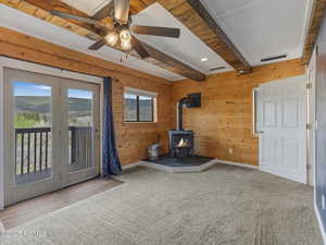 Unfurnished living room featuring wood walls, beamed ceiling, a wood stove, ceiling fan, and carpet floors