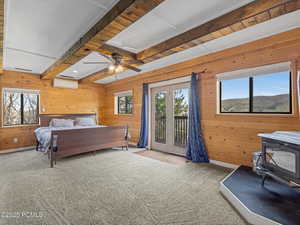 Bedroom with wooden walls, beamed ceiling, access to exterior, light colored carpet, and a wood stove