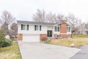 Raised ranch featuring a front lawn, an attached garage, concrete driveway, brick siding, and a chimney