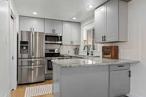 Kitchen with light stone counters, stainless steel appliances, a peninsula, gray cabinets, and light wood-style flooring