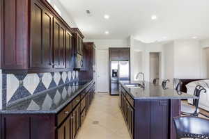 Kitchen with a breakfast bar, dark stone countertops, an island with sink, dark wood finish cabinets, and stainless steel appliances