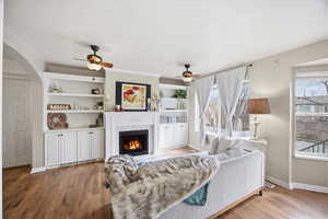 Living room featuring light wood-style floors, a ceiling fan, and a warm lit fireplace