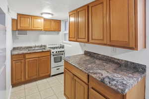 Kitchen featuring white gas stove, wood finish cabinets, a textured ceiling, dark countertops, and light tile patterned flooring
