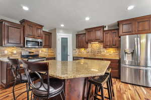 Kitchen featuring stainless steel appliances, light stone countertops, a kitchen breakfast bar, light wood-type flooring, and dark wood finish cabinetry