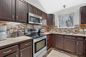 Kitchen featuring stainless steel appliances, dark wood finish cabinets, vaulted ceiling, light countertops, and tasteful backsplash