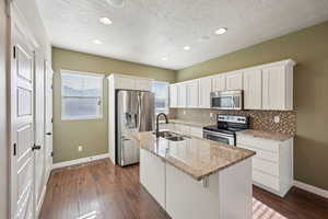 Kitchen featuring white cabinetry, stainless steel appliances, light stone counters, a center island with sink, and dark wood finished floors