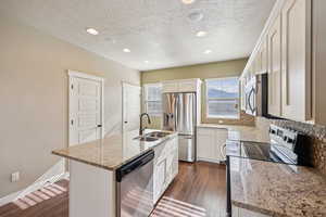 Kitchen with an island with sink, stainless steel appliances, light stone counters, dark wood finished floors, and white cabinetry