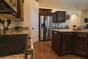 Kitchen featuring dark wood finish cabinetry, stainless steel refrigerator with ice dispenser, arched walkways, and light stone counters
