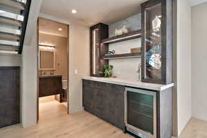 Indoor bar featuring wine cooler, open shelves, dark wood finish cabinetry, light wood finished floors, and glass insert cabinets