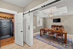 Dining room with light wood-style floors, a barn door, a raised ceiling, a chandelier, and brick wall