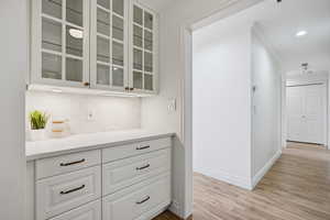 Bar featuring white cabinetry, glass fronted cabinets, light wood-style flooring, tasteful backsplash, and crown molding