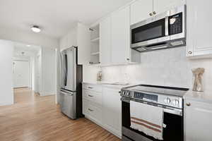 Kitchen with stainless steel appliances, white cabinets, open shelves, light wood finished floors, and decorative backsplash