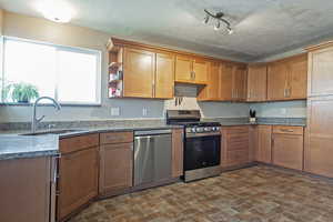 Kitchen featuring stainless steel appliances, wood finish cabinetry, brick patterned flooring, dark stone countertops, and a textured ceiling