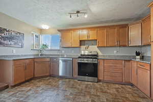 Kitchen with stainless steel appliances, light stone countertops, wood finish cabinetry, brick patterned floors, and a textured ceiling