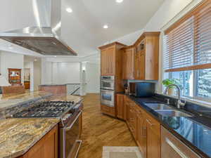 Kitchen with dark stone counters, stainless steel appliances, island range hood, wood finish cabinets, and light wood-style floors