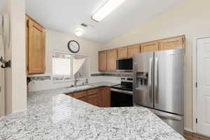 Kitchen featuring stainless steel appliances, backsplash, vaulted ceiling, light stone counters, and wood finish cabinets