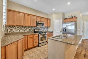 Kitchen with stainless steel appliances, light stone counters, a center island with sink, wood finish cabinetry, and tasteful backsplash