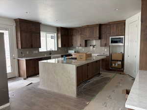 Kitchen with light stone countertops, a kitchen island with sink, stainless steel microwave, backsplash, and recessed lighting