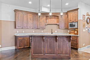 Kitchen with an island with sink, stainless steel microwave, light stone countertops, and wood finish cabinetry