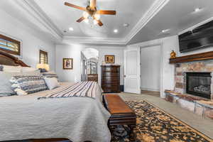 Carpeted bedroom with arched walkways, a stone fireplace, crown molding, a tray ceiling, and a ceiling fan