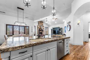 Kitchen with a stone fireplace, a center island with sink, a ceiling fan, dark stone counters, and stainless steel dishwasher