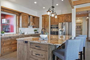 Kitchen with wood finish cabinets, backsplash, light stone countertops, a center island with sink, and stainless steel appliances
