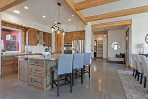 Kitchen with beamed ceiling, wood finish cabinetry, light stone countertops, decorative backsplash, and an island with sink