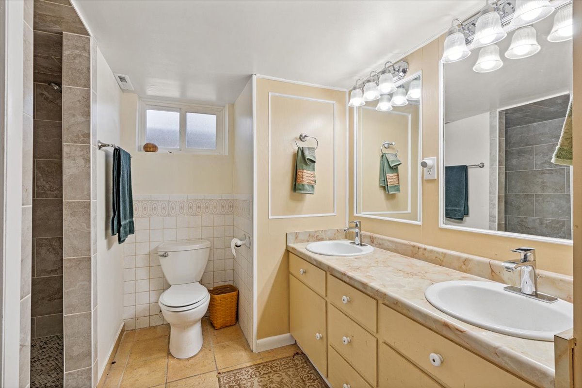 Bathroom featuring double vanity, tile walls, tiled shower, and a wainscoted wall