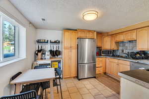 Kitchen with light wood finish cabinetry, stainless steel appliances, decorative backsplash, dark countertops, and light tile patterned floors