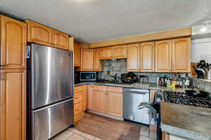 Kitchen featuring stainless steel appliances, light wood finish cabinetry, light tile patterned flooring, a textured ceiling, and decorative backsplash