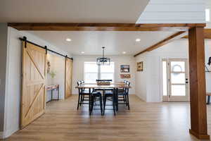 Dining area featuring a barn door, light wood-style flooring, wooden walls, suspended lighting, and beamed ceiling