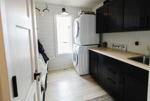 Laundry room with stacked washing machine and dryer, light wood-style flooring, cabinet space, and wooden walls